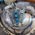 A silver butterfly pendant with four round turquoise stones, displayed on a natural clam shell with rocks in the background.