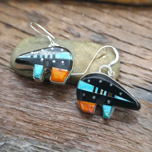 A pair of handmade geometric inlay  bear earrings resting on a small stone over a rustic wooden surface. The earrings feature a black stone base with turquoise blue, orange, and silver-colored inlay details.