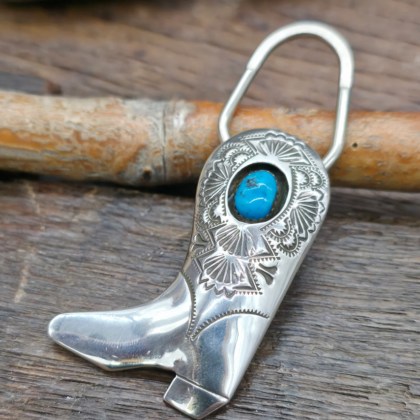 A silver cowboy boot keychain with an engraved floral pattern and a round turquoise stone set near the top, photographed on a rustic wooden surface with a branch behind i