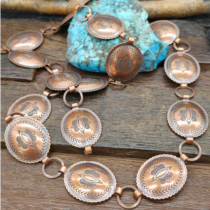 A solid copper Southwestern-style necklace with oval engraved pendants and circular links, displayed on a rustic wooden surface with a turquoise stone and driftwood.