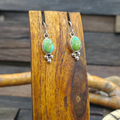 A pair of sterling silver drop earrings with oval green turquoise stones and small bead drops, displayed on a wooden stand against a rustic wooden background.