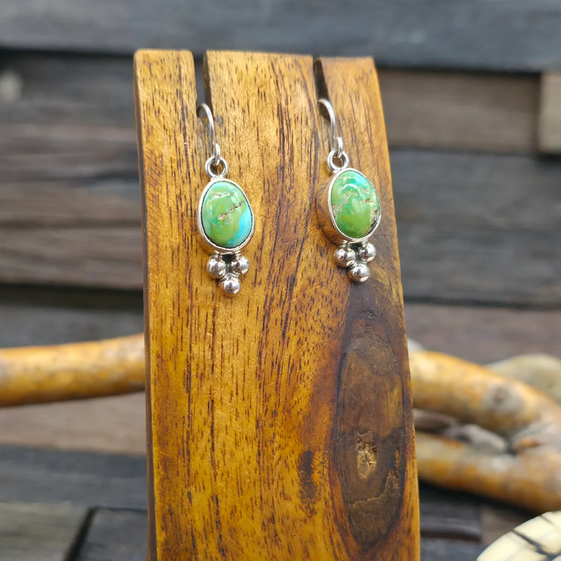 A pair of sterling silver drop earrings with oval green turquoise stones and small bead drops, displayed on a wooden stand against a rustic wooden background.