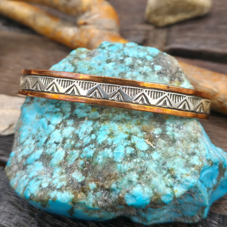 A silver geometric-patterned cuff bracelet with copper  edges, displayed on a bright turquoise stone over a dark wooden surface.