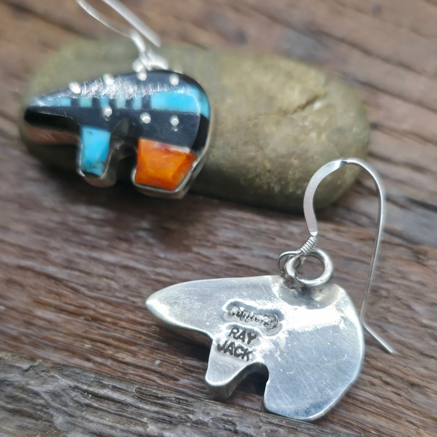 A pair of handmade silver bear dangle earrings with colorful inlay stones, displayed on a rustic wooden surface with a smooth stone prop. One earring is shown from the front, featuring turquoise, orange, and black inlay, while the other is shown from the back, revealing the sterling silver cast and the engraved brand name 'Ray Mack'.